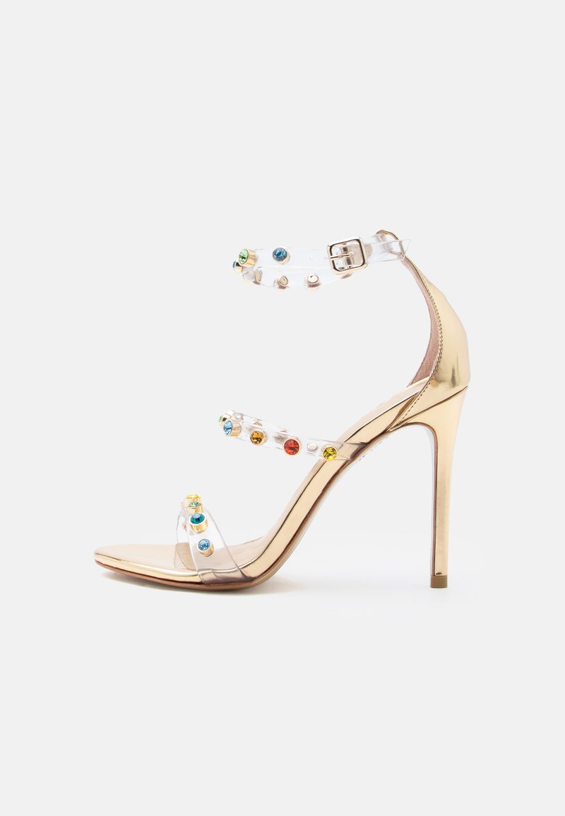 Χρυσό πέδιλο υψηλού τακουνιού με τρεις διαφανείς λουρίδες διακοσμημένες με πολύχρωμους πολύτιμους λίθους και κούμπωμα στον αστράγαλο.