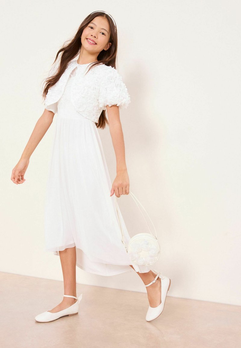 Niña joven con un vestido blanco y una torera texturizada, sosteniendo un bolso blanco redondo, con zapatos blancos de tiras en el tobillo, de pie sobre un suelo claro.