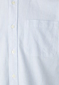 Camisa de botones a rayas azul claro y blanca, hecha de tela tejida, que cuenta con un bolsillo de parche en el lado izquierdo y botones de plástico.