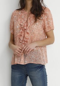 Peach floral blouse with ruffled front detail, short sleeves, and button closures, paired with blue jeans. Sheer fabric with a delicate pattern.