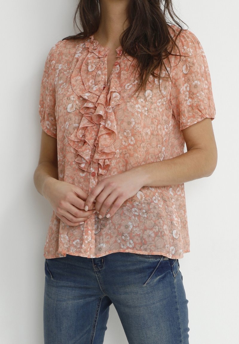 Peach floral blouse with ruffled front detail, short sleeves, and button closures, paired with blue jeans. Sheer fabric with a delicate pattern.