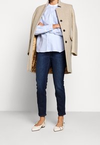 Manteau en laine beige avec boutons noirs, chemise boutonnée bleu clair, jean denim foncé, et ballerines pointues couleur crème. Design simple et minimaliste.