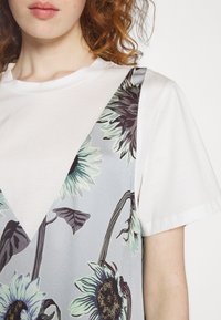 Jeune personne aux cheveux roux bouclés porte une chemise blanche à manches courtes sous une robe grise avec un grand imprimé de tournesol vert clair et noir.