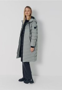 Vadderad grå kappa med huva, lång silhuett, elastiska mudd och sidfickor. Har svart accent på ärmen och en slät, blanka yta.