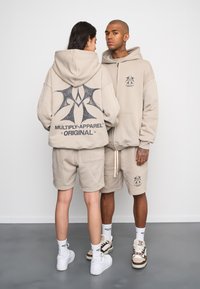 Felpe con cappuccio beige e pantaloni corti con loghi frontali e posteriori. Texture liscia, vestibilità rilassata, pantaloni con coulisse e sneakers bianche.