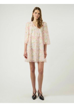 Kobieta stojąca w pastelowej sukience w kwiaty do kolan z rękawami do ćwiartki oraz czarnych czółenkach na obcasie z paskiem z tyłu.