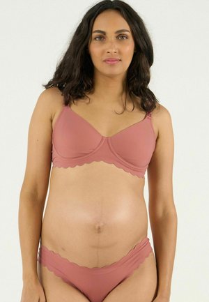 Femme enceinte aux longs cheveux foncés portant un soutien-gorge et des sous-vêtements rose assortis avec des bords festonnés, debout devant un fond uni.