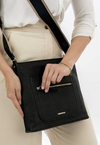 Black leather crossbody bag with textured surface, accent zip pocket, and gold branding. Features a wide, adjustable strap.