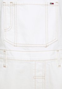 Wit denim kledingstuk met beige stiksels, voorzien van een grote vierkante achterzak en ceintuurlussen, met een klein rood-wit-blauw label.