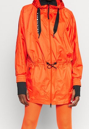 Personne portant une veste à capuche orange vif avec des cordons noirs et des leggings en résille orange, debout devant un fond clair et uni.