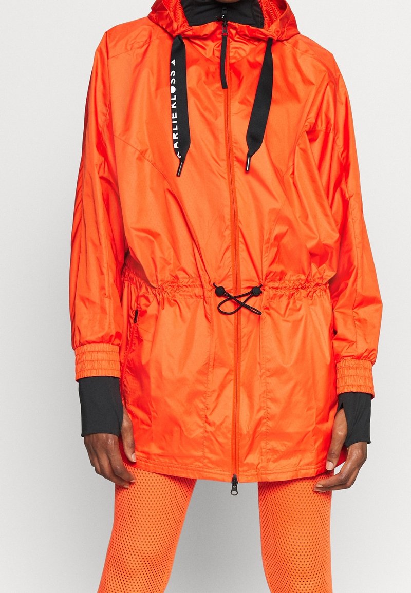 Personne portant une veste à capuche orange vif avec des cordons noirs et des leggings en résille orange, debout devant un fond clair et uni.