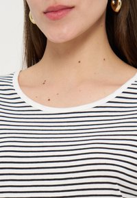 Κάτω μέρος προσώπου και λαιμός γυναίκας με σπίλους, που φοράει χρυσούς κρίκους στα αυτιά και άσπρο πουκάμισο με λεπτές μαύρες οριζόντιες ρίγες.