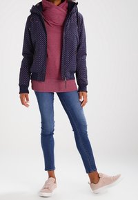 Persona che indossa una giacca blu navy con pois sopra una felpa bordeaux, jeans skinny blu e sneakers rosa chiaro, in piedi con una gamba leggermente sollevata.