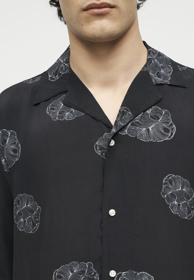 Κοντινό πλάνο ενός ατόμου που φοράει μαύρο πουκάμισο με κουμπιά και ανοιχτό γιακά, με λευκά floral σχέδια.