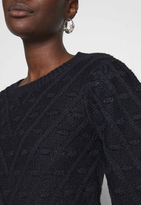 Marinblå stickad tröja med ett texturerat mönster och upphöjda detaljer samt vid halsringning. Kompletteras av en stor cirkulär silverörhänge.