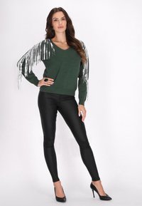 Pulover verde cu mâneci cu franjuri argintii, design în formă de V. Asortat cu pantaloni negri ajustați și tocuri negre înalte. Textură netedă în ansamblu.