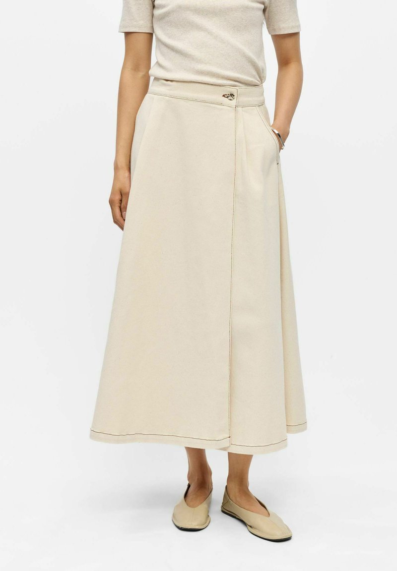 Femme portant une jupe midi beige portefeuille avec poche latérale et un haut beige assorti ainsi que des chaussures, sur fond blanc uni.