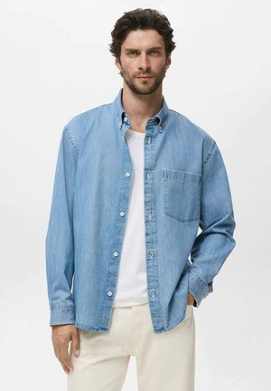 Light blue denim shirt with button-up front, pointed collar, and single chest pocket. Worn over a white t-shirt, paired with light pants.