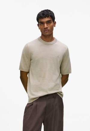 Mand iført en beige kortærmet sweater med et lille logo og mørkebrune plisserede bukser, stående med hænderne bag ryggen.