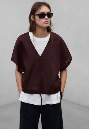 Młoda osoba w ciemnych, dużych okularach przeciwsłonecznych, luźnej bordowej bluzce z dekoltem w serek na białej koszuli, czarne spodnie, stojąca na szarym tle.