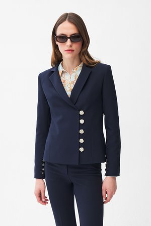 Donna che indossa un blazer navy su misura con bottoni decorativi argentati, una camicetta floreale, pantaloni coordinati navy e occhiali da sole ovali scuri.