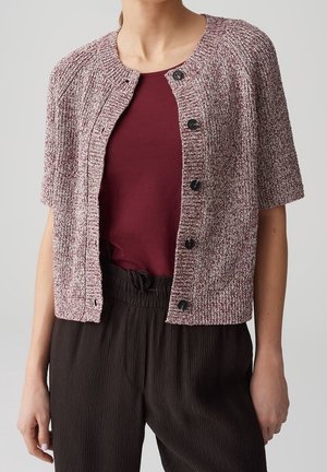 Femme portant un haut bordeaux sous un cardigan à manches courtes texturé rose et blanc boutonné, avec un pantalon ample marron foncé.