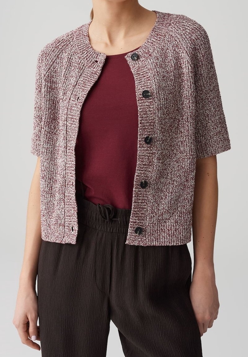 Femme portant un haut bordeaux sous un cardigan à manches courtes texturé rose et blanc boutonné, avec un pantalon ample marron foncé.
