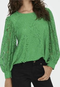 Femme portant un chemisier à manches longues en dentelle florale verte avec des manches ballon, associé à un pantalon noir, main reposant dans la poche.