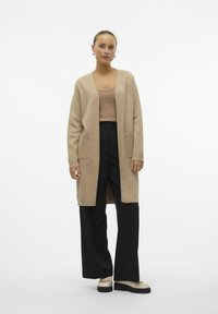 Bež pletena jopica z dolgimi rokavi, odprta spredaj, s stranskimi žepi, nosi se čez gole majice in črne široke hlače, v kombinaciji s svetlimi čevlji.