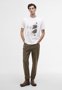 Man standing with hands in pockets wearing white t-shirt with cow graphic, olive pants, and brown boots on plain background.