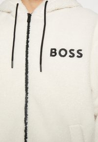 Weißer Fleece-Hoodie mit schwarzem Reißverschluss und Kordelzug, mit "BOSS" in Schwarz auf der linken Brustseite gestickt.