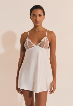 Vrouw die een witte satijnen slipjurk draagt met doorzichtige cups afgewerkt met kant en dunne schouderbandjes, staand tegen een beige achtergrond.