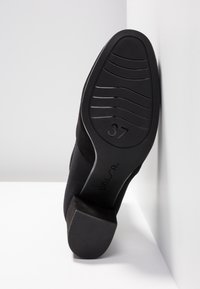 Semelle de chaussure à talon noire taille 37, avec un motif de bande de roulement incurvé et le logo "UNISA", positionnée contre un mur d'angle blanc.