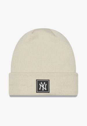 Gorro de punto cremoso con puño doblado, que presenta un parche cuadrado negro con un logo blanco. Textura suave, patrón acanalado.