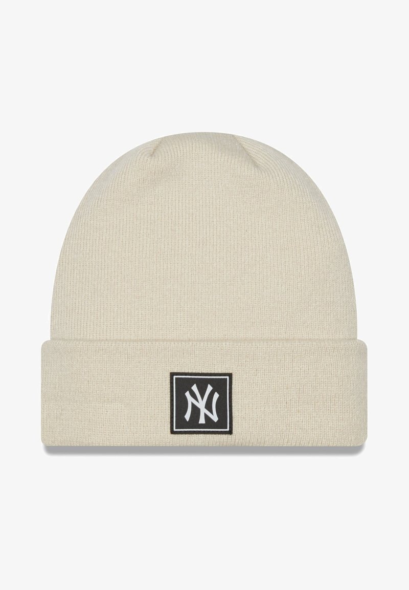 Cream knitted beanie with a folded cuff, featuring a black square patch displaying a white logo. Soft texture, ribbed pattern.