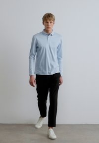 Γαλάζιο μακρυμάνικο πόλο πουκάμισο, μαύρο εφαρμοστό παντελόνι και λευκά αθλητικά παπούτσια. Το μοντέλο στέκεται σε γκρι πάτωμα με λευκό φόντο.