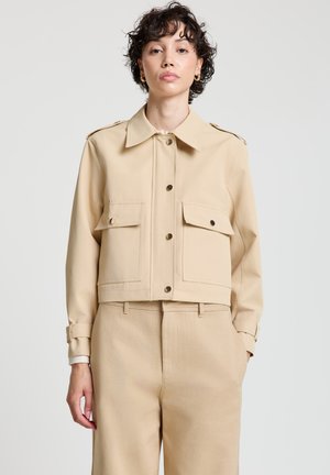 Beige cropped jacket with two front pockets, button closure, and epaulets. Smooth fabric and cuffed sleeves. Paired with beige trousers.