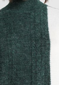 Pull à col roulé en tricot vert foncé et blanc, présentant un motif texturé sur la partie verte et un tissu blanc lisse.