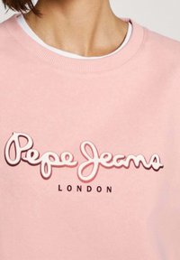 Sudadera rosa claro con textura suave; presenta el logo "Pepe Jeans" en blanco y rosa oscuro, y "LONDON" en negro debajo.