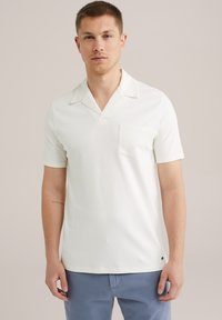 Polo alb cu mânecă scurtă dintr-un material moale, având guler în V, și un singur buzunar pe piept. Branding subtil la tiv.