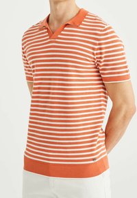 Man som bär en kortärmad orange och vit randig polotröja med krage, i kombination med vita byxor, stående med händerna bakom ryggen.