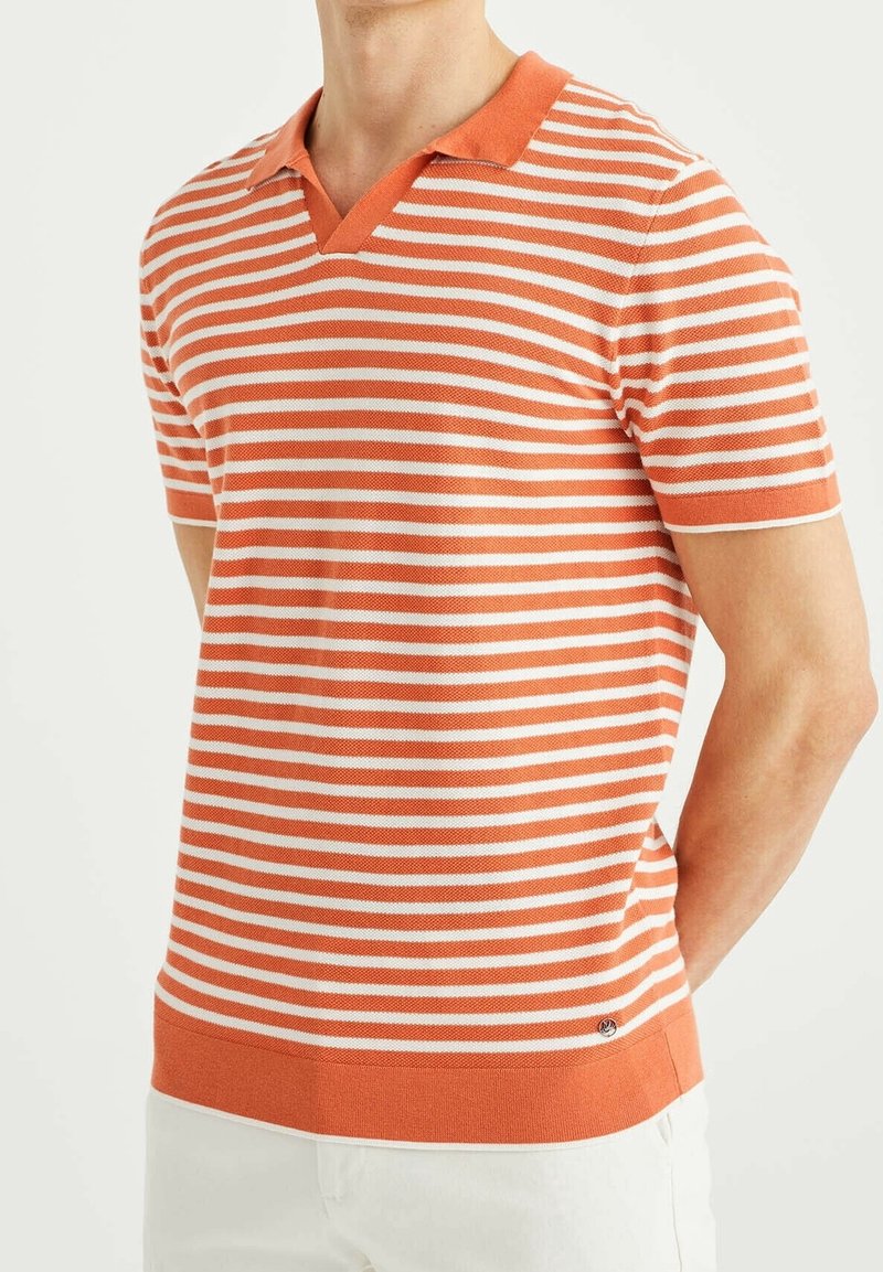 Man som bär en kortärmad orange och vit randig polotröja med krage, i kombination med vita byxor, stående med händerna bakom ryggen.