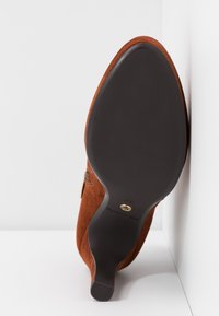 Chaussure à talon en daim marron positionnée talon vers le haut, montrant une semelle noire lisse avec la taille 37 et un détail doré près de la voûte plantaire.