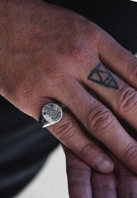 Anello sigillo in argento con incisioni floreali intricate, forma arrotondata, indossato su una mano con visibile texture della pelle e tatuaggi.