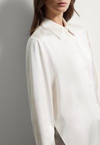 Una donna in camicia a maniche lunghe, bottone, di colore crema con colletto a punta, in piedi contro uno sfondo grigio uniforme.