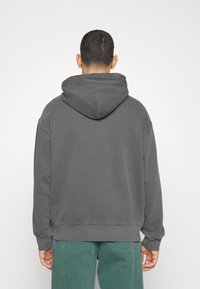 Uomo di spalle che indossa una felpa grigia scura larga con cappuccio e pantaloni verde teal su uno sfondo bianco uniforme.