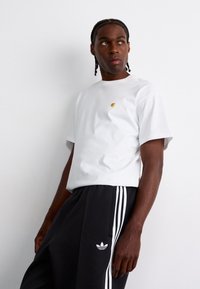 White short-sleeve t-shirt with a small yellow logo, paired with dark gray sweatpants featuring white three-stripe accents and logo.