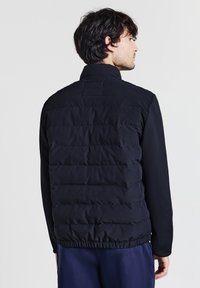 Homme avec des cheveux ondulés foncés portant une veste matelassée noire avec une ceinture élastique et un pantalon bleu marine, de profil contre un fond uni.