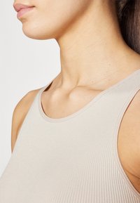 Nøytral beige ribbet tanktopp med høy hals og tykke stropper, som har en glatt tekstur og subtile sømdetaljer.
