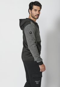Sudadera de hombre en gris y negro. Presenta un cuerpo de textura suave, mangas en contraste y una capucha con cordón. Incluye un logo redondo en la manga.